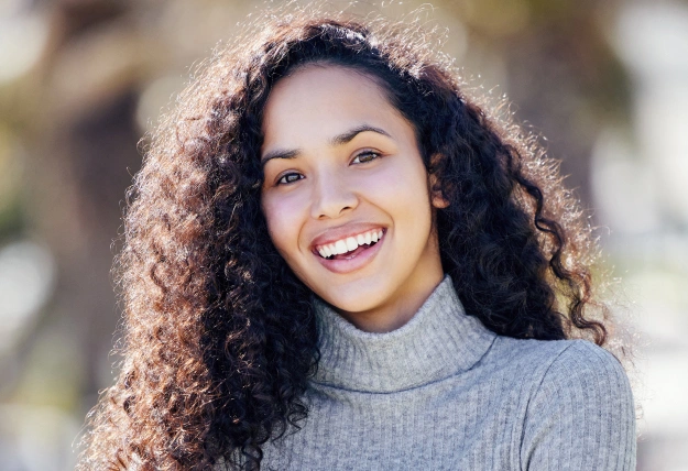 Young woman with curly hair smiling outdoors wearing gray turtleneck sweater.