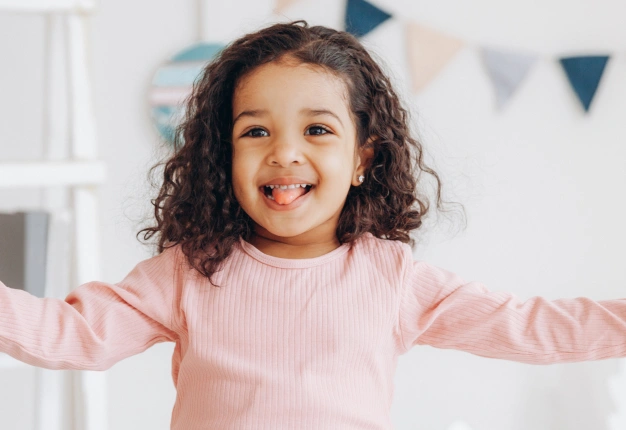 Happy young child with curly hair smiling with arms outstretched in bright, cheerful indoor setting.