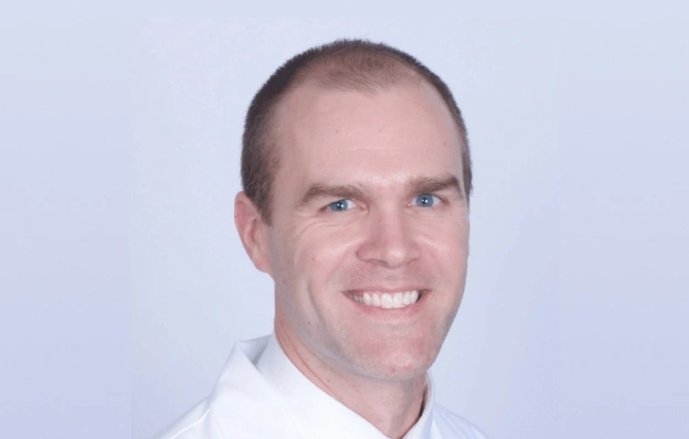 Professional headshot of smiling male dentist wearing white coat against light blue background.