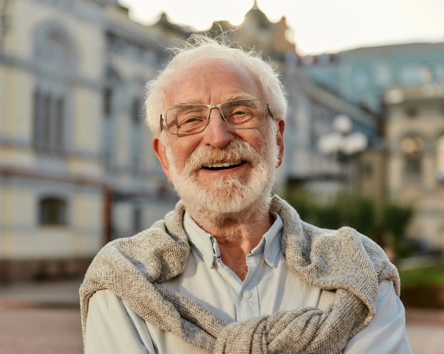 Smiling older man with glasses wearing vest outdoors in front of historic buildings.