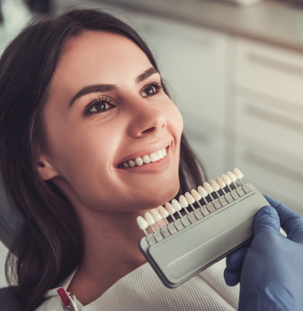 Female dental professional holding tooth shade guide against face, smiling in clinical setting.