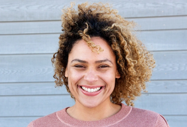 Smiling woman with curly hair wearing mauve shirt against blue wooden wall backdrop.