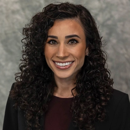 Professional headshot of smiling woman with curly dark hair in dental office setting.