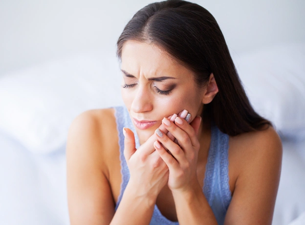 Woman holding cheek in pain, sitting in dental office chair, experiencing tooth discomfort.