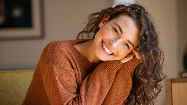 Smiling woman in brown sweater relaxing on green couch in modern dental office setting.