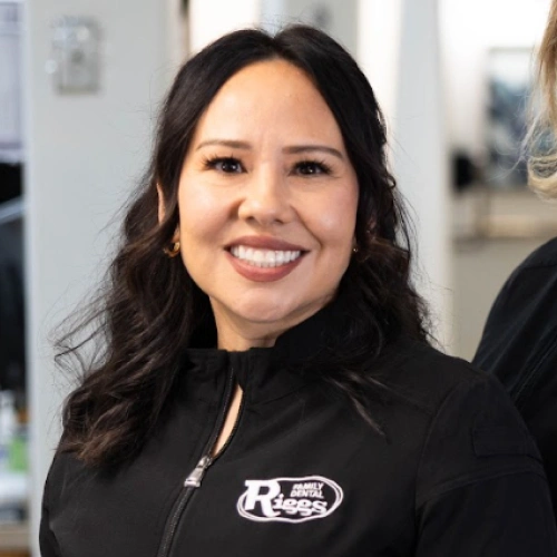 Woman smiling at camera wearing black Riggs Family Dental branded jacket in dental office setting.