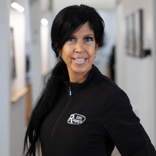 Professional woman smiling in dental office hallway wearing black Riggs Family Dental branded jacket.
