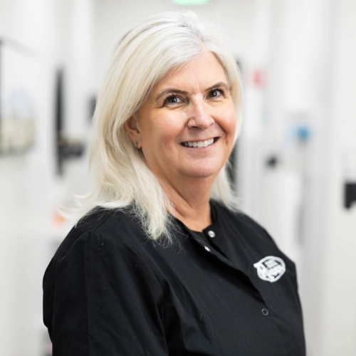 Professional woman with white hair smiling in modern dental office hallway wearing black uniform.