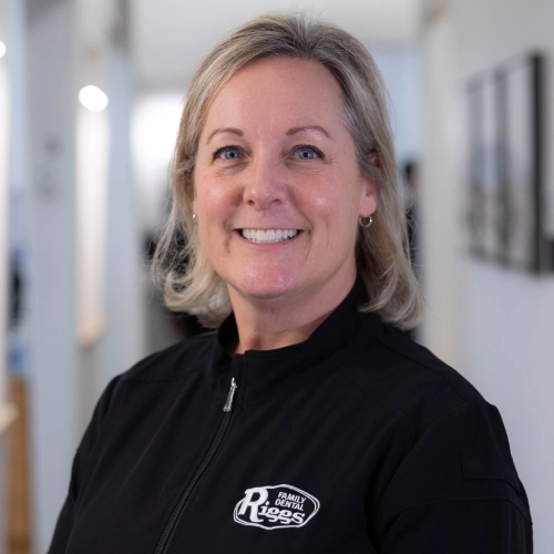 Woman smiling at camera in dental office hallway wearing black Riggs Family Dental jacket.