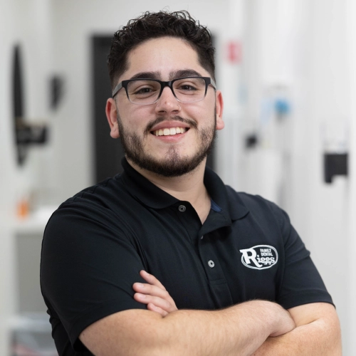 Young man wearing glasses and black polo shirt with dental practice logo, smiling with arms crossed in dental office setting.