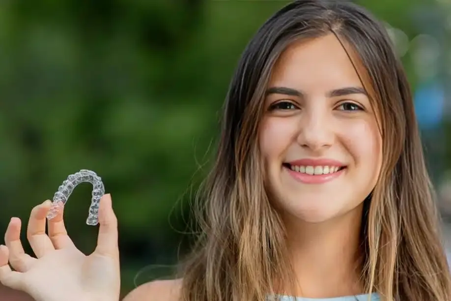 Young woman smiling outdoors holding clear dental aligner with green background