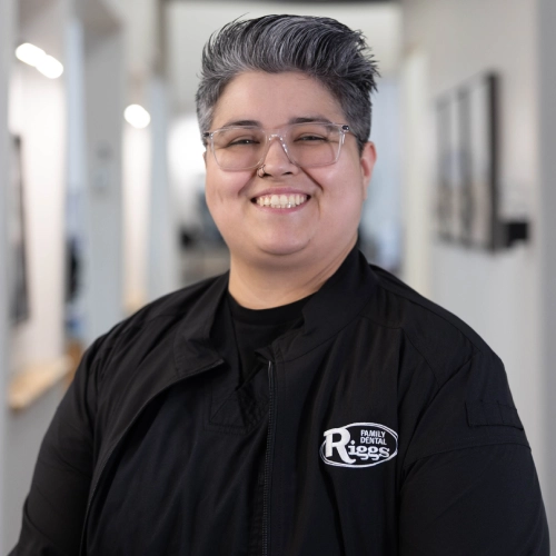 Smiling dental staff member wearing glasses and black Riggs Family Dental uniform in clinical hallway.