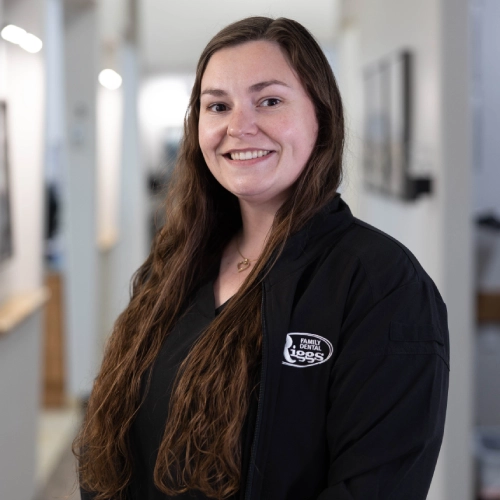Young woman in black Riggs Family Dental polo smiling in modern dental office hallway.