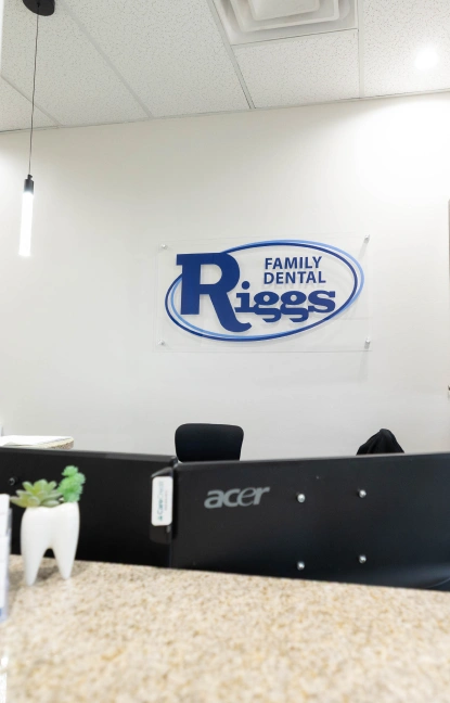 Riggs Family Dental reception desk with logo on white wall and black counter.