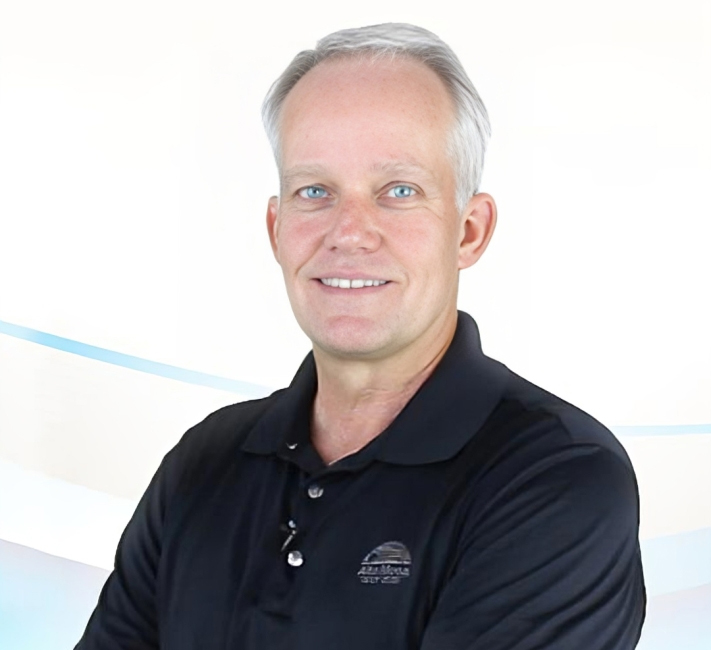 Professional headshot of male dentist smiling at camera wearing black polo shirt against white background.
